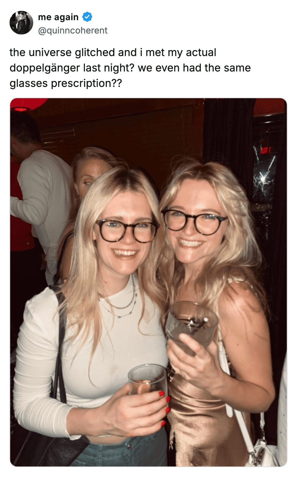 Two blonde women wearing identical black-rimmed glasses and smiling together at a night event.