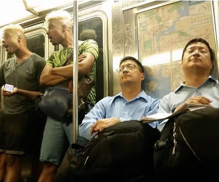 Two pairs of doppelgängers sitting and standing near each other on a crowded subway train.