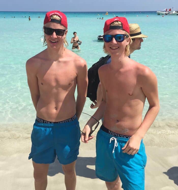 Two teenage boys with blonde hair, red hats, and blue swim trunks standing on a beach.
