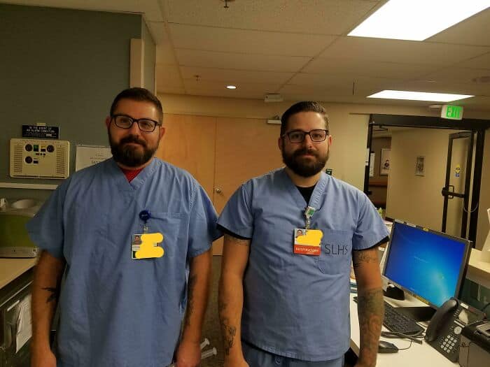 Two male healthcare workers in blue scrubs with dark hair, beards, and glasses standing together.