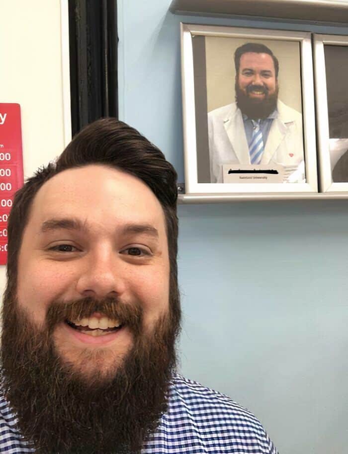 A bearded man taking a selfie next to a framed professional portrait of his lookalike.