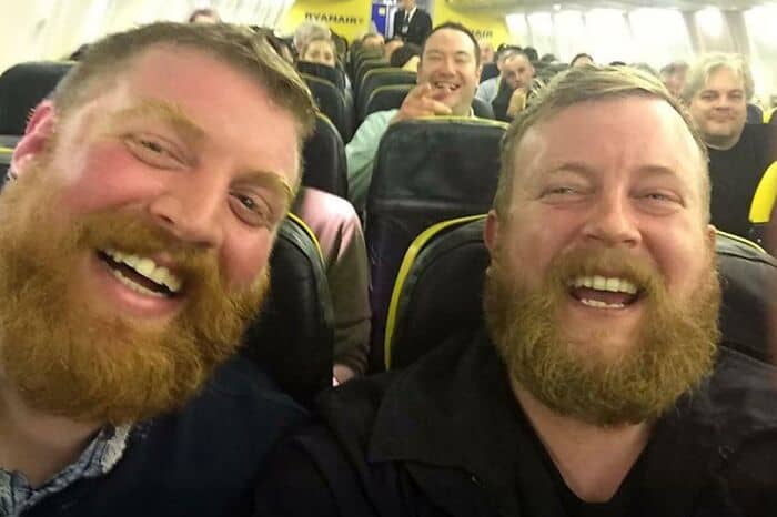 Two men with identical ginger beards laughing hysterically while sitting next to each other on a plane.