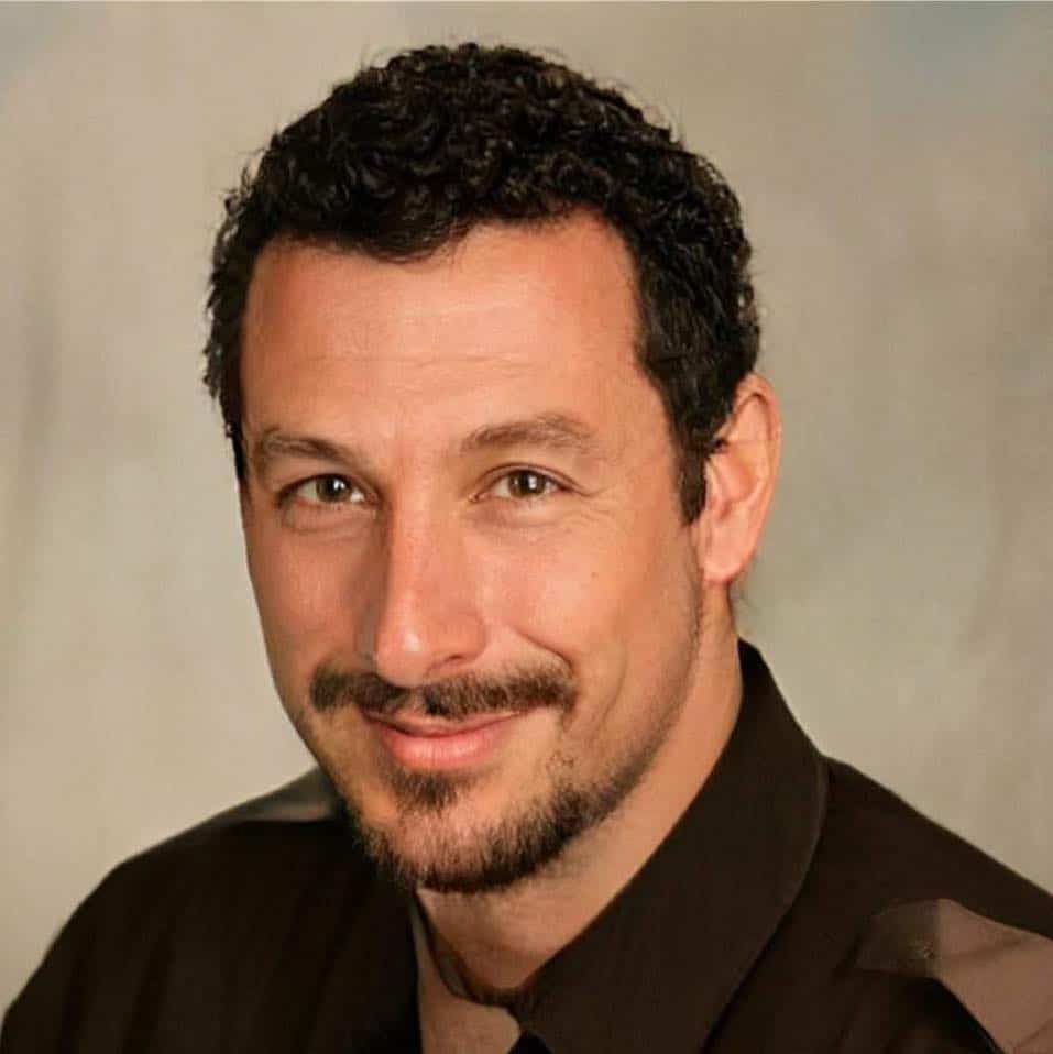 Close up portrait of a man with curly hair and a goatee who looks like Adam Sandler.
