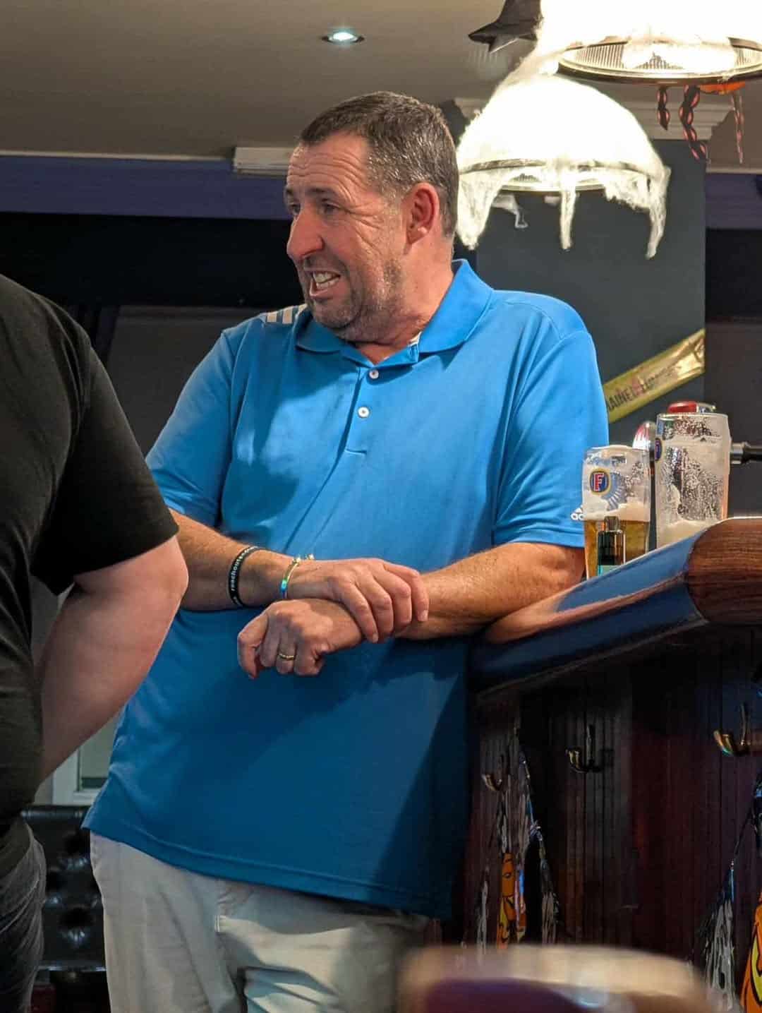 An older man in a blue polo shirt leaning against a bar with Sandler's distinct profile.