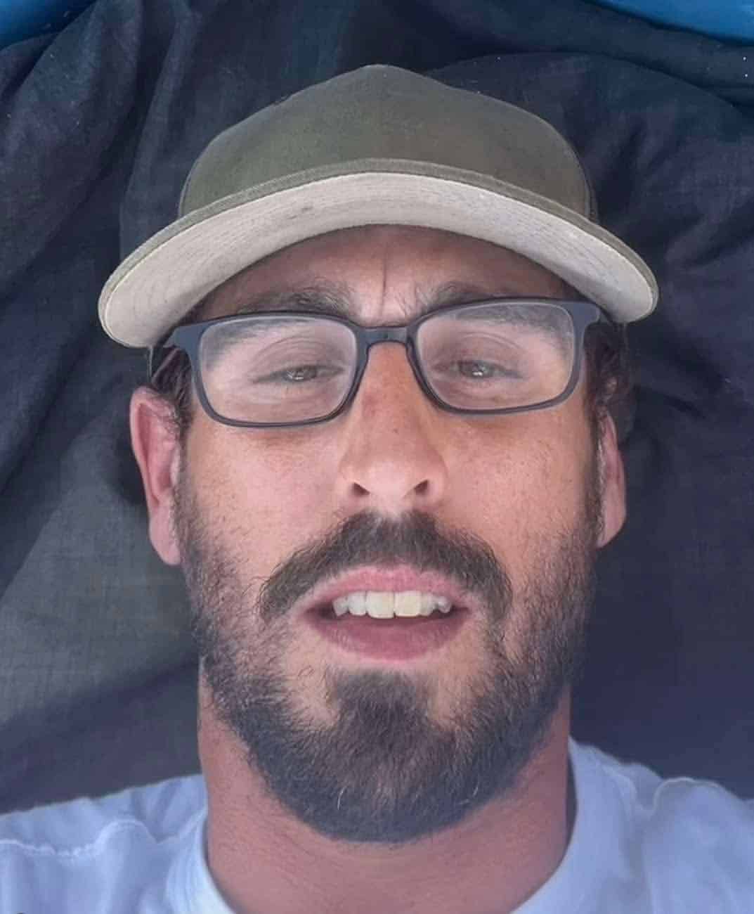 Low angle selfie of a man wearing glasses and a baseball cap with Sandler-esque facial features.
