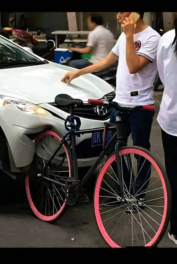 A black bicycle with pink rims leaning against a white car with a large front dent.