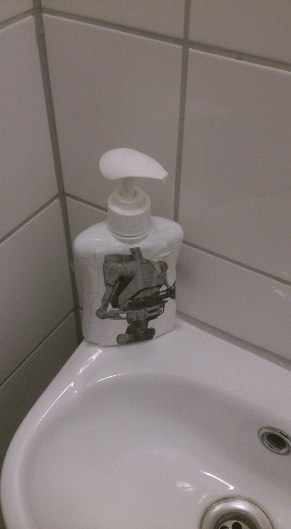 real-life star wars meme showing a printed picture of a B1 Battle Droid taped perfectly onto a white bathroom soap dispenser, making the pump look exactly like the droid's long head.