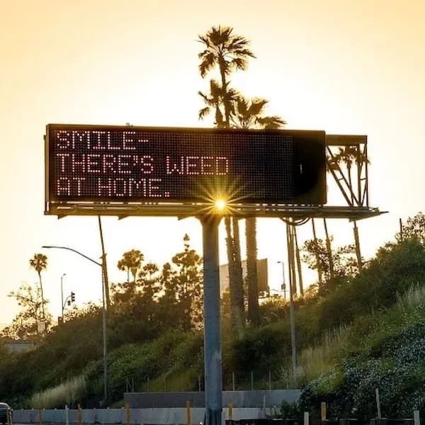 wholesome and motivational stoner meme featuring a massive digital highway billboard glowing beautifully at sunset with the incredibly comforting message: "Smile - there's weed at home," an iconic weed meme.