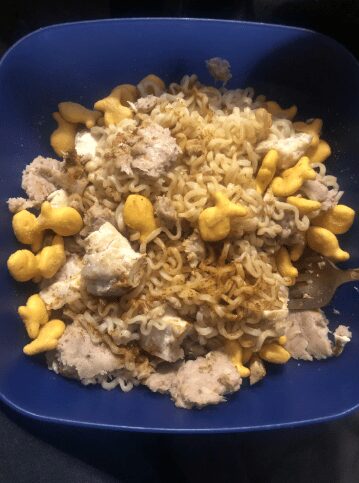 Blue bowl containing instant ramen noodles topped with chunks of tuna and yellow Goldfish crackers.