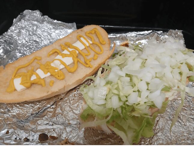 Hoagie roll topped with a single slice of cheese, mustard, shredded lettuce, and diced white onions.