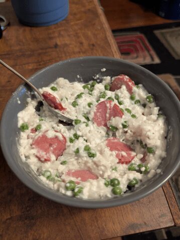 Gray bowl filled with a creamy mixture of white rice, green peas, and sliced hot dogs.