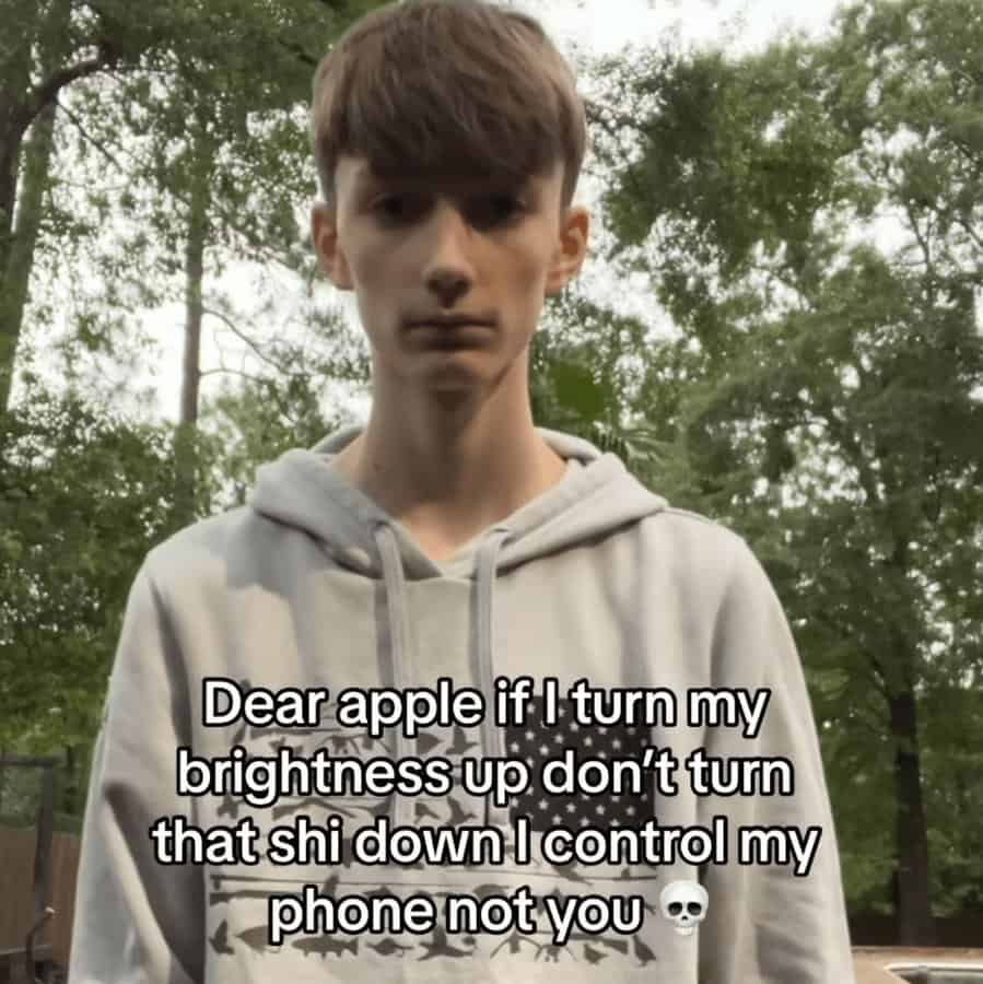 defiant unhinged screenshot showing a thin teenager in a grey hoodie. The text is an open letter to Apple, aggressively asserting dominance over his device’s settings: "Dear apple if I turn my brightness up don’t turn that shi down I control my phone not you."