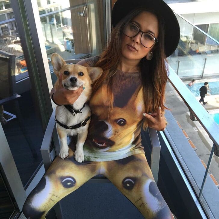 A woman sits on a balcony holding a real Corgi while wearing a matching t-shirt and leggings set featuring a massive, wide-eyed, hyper-realistic print of a Corgi's face, creating a dizzying and surreal meta-fashion moment.