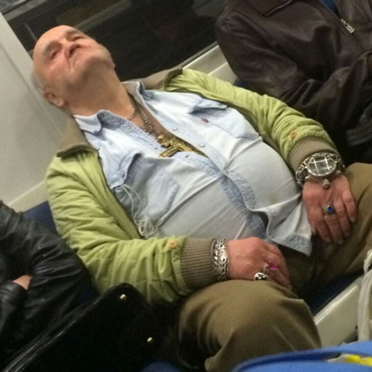 Capturing the "final boss of jewelry," an older man sleeps on a train wearing an excessive collection of heavy metal bracelets, an oversized bulky watch, and multiple large gemstone rings over a simple green bomber jacket.
