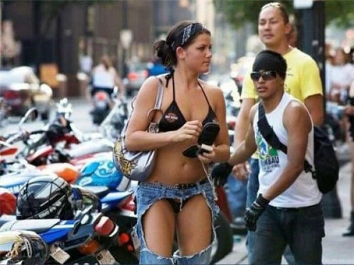 A bold summer look on a city street features a woman in a black bikini top and "chapped" denim jeans with the thighs entirely removed, exposing her legs from the waist down to the shins while maintaining the denim cuffs.