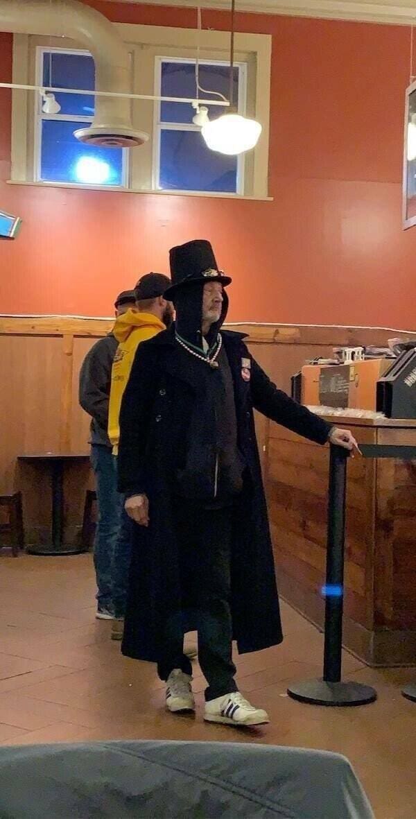 A chaotic streetwear ensemble consisting of a tall black top hat, a long trench coat worn over a hoodie, a festive beaded bell necklace, and basic white sneakers.