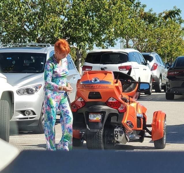 Extreme coordination featuring a woman with bright orange hair and a vibrant floral tracksuit standing next to her perfectly color-matched orange three-wheeled motorcycle