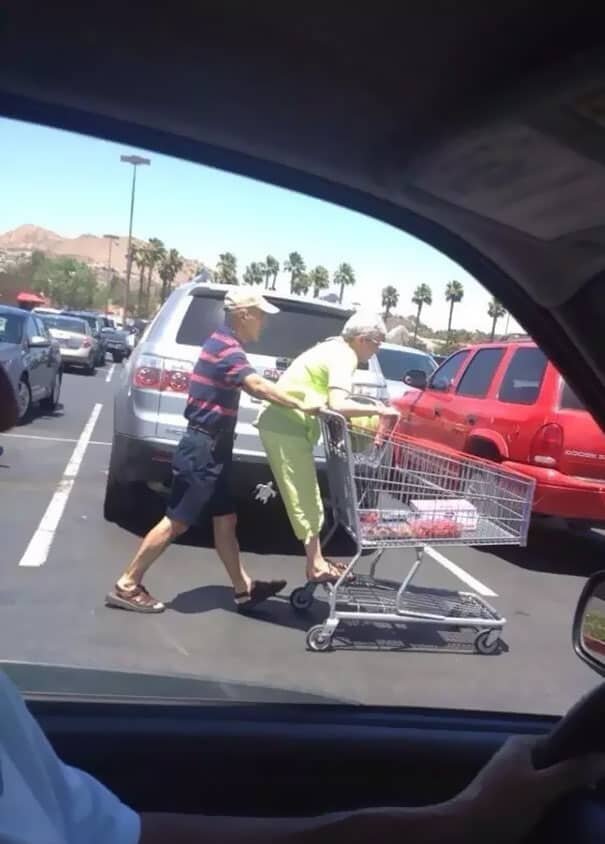 Elderly woman standing on the bottom rail of a shopping cart being pushed by a man.