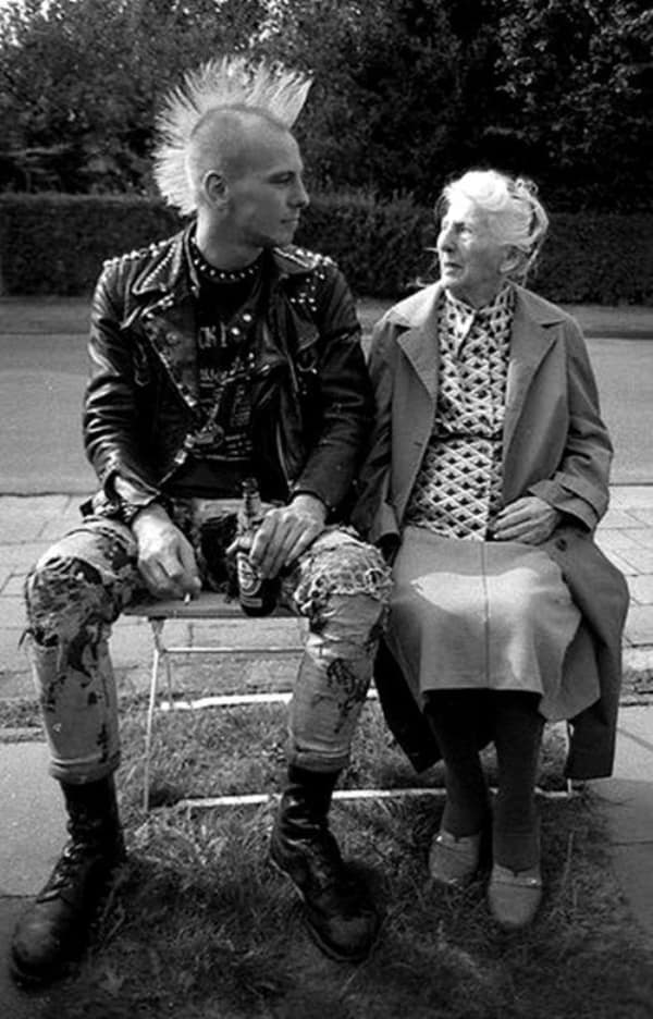 powerful wholesome image captured in black and white, showing a punk with a giant mohawk and an elderly woman sharing a quiet, respectful, and friendly moment while sitting together on a bench.
