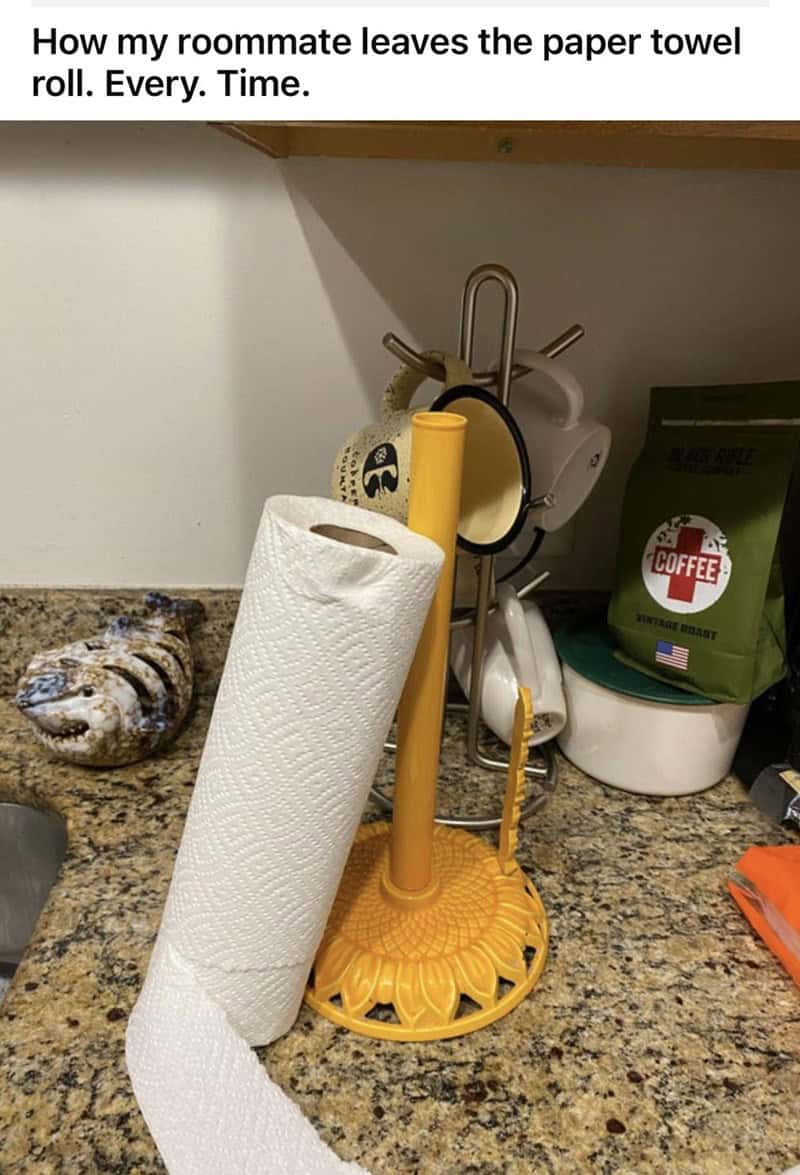 A low-effort fail from one of the worst roommates. A yellow paper towel stand sits on a granite counter, but the roll is simply leaning against the spindle diagonally—and unravelling—rather than being slid onto it properly.