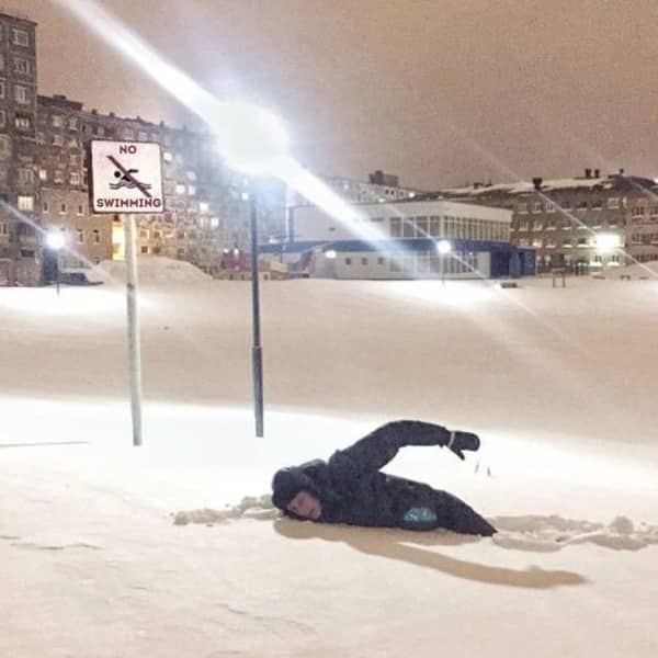 Person lying in deep snow next to a no swimming sign in a city.