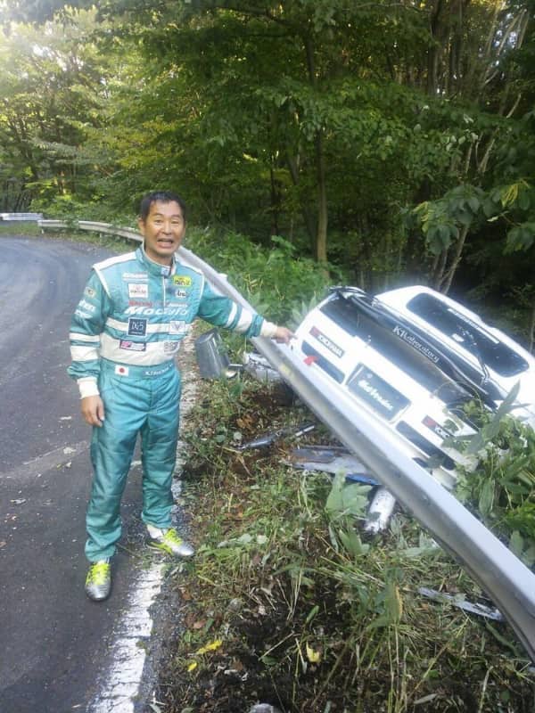 Race car driver in green suit pointing at a car crashed into a guardrail.
