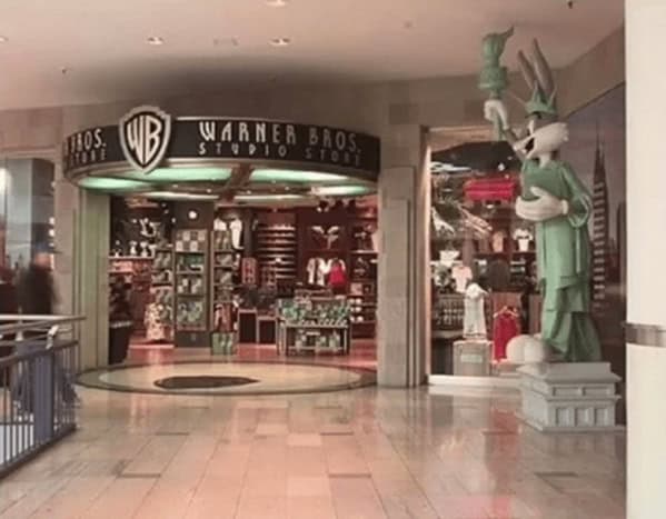 High-energy 90s mall glamour shots featuring the entrance to a Warner Bros. Studio Store. A giant Bugs Bunny statue dressed as the Statue of Liberty stands guard outside the dark wood and neon-lit storefront, perfectly capturing the era's mix of retail and entertainment