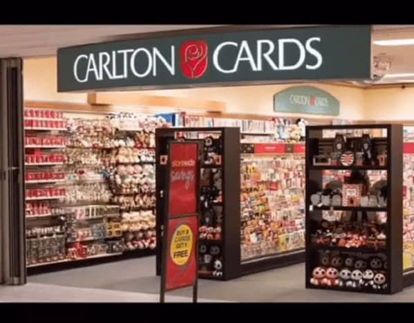 A cozy 90s mall glamour shots capture of a Carlton Cards shop. The well-lit entrance reveals shelves overflowing with greeting cards and 90s-era stuffed animals, all under a clean, backlit sign featuring the brand's iconic red rose.