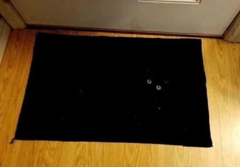 A "void" accidental camouflage photo featuring a black cat lying on a rectangular black doormat. The cat is completely invisible against the dark background except for two round, glowing yellow eyes staring directly at the camera.