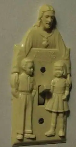 A vintage beige plastic light switch cover featuring a 3D relief of Jesus Christ with his arms around two children, with the toggle switch protruding from a highly unfortunate anatomical position.