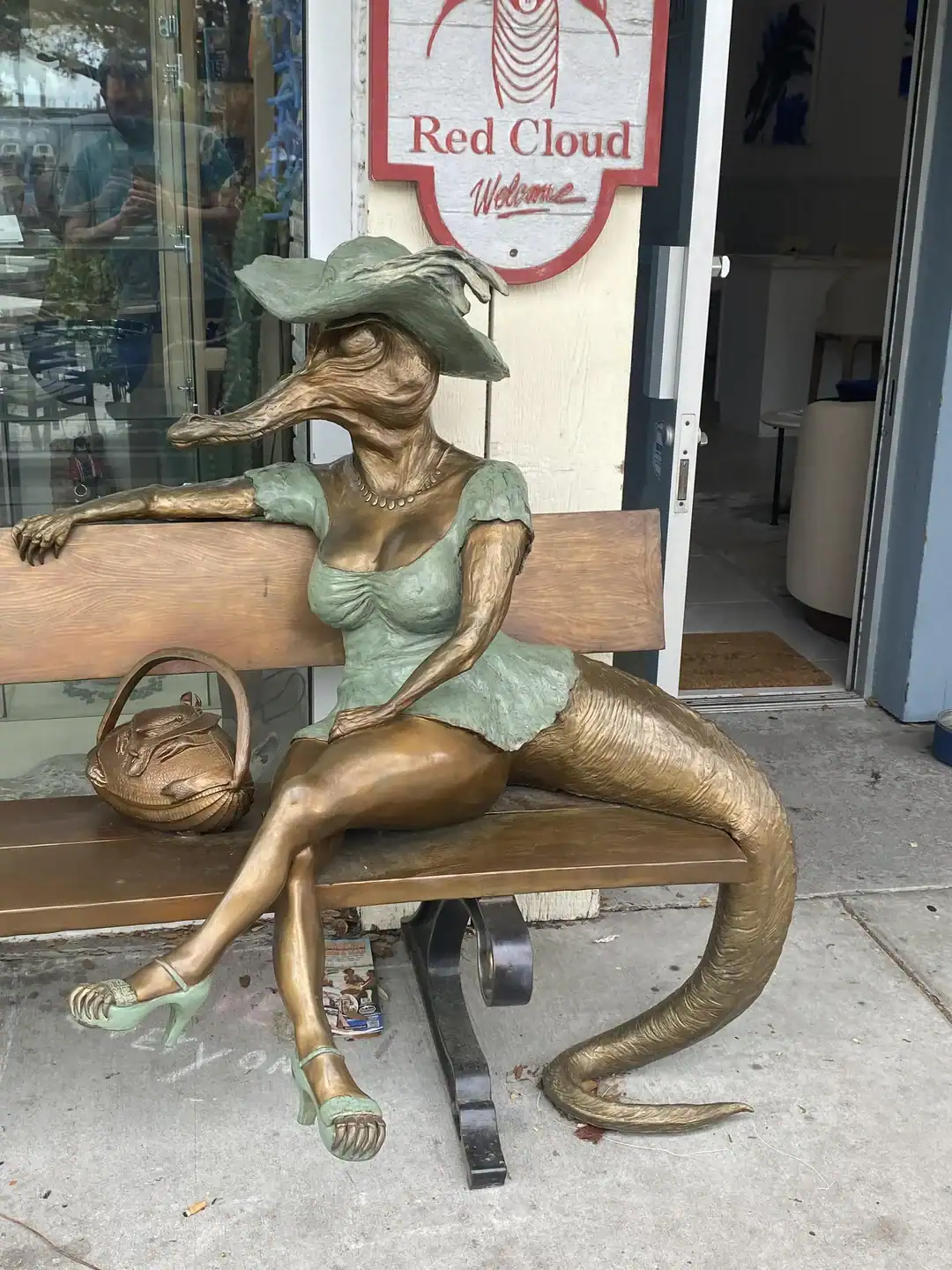 bizarre bronze sculpture of a humanoid crocodile woman sitting on a park bench. She is dressed in a green sun hat, a matching top, and high heels, with a thick reptilian tail curling off the side of the seat.