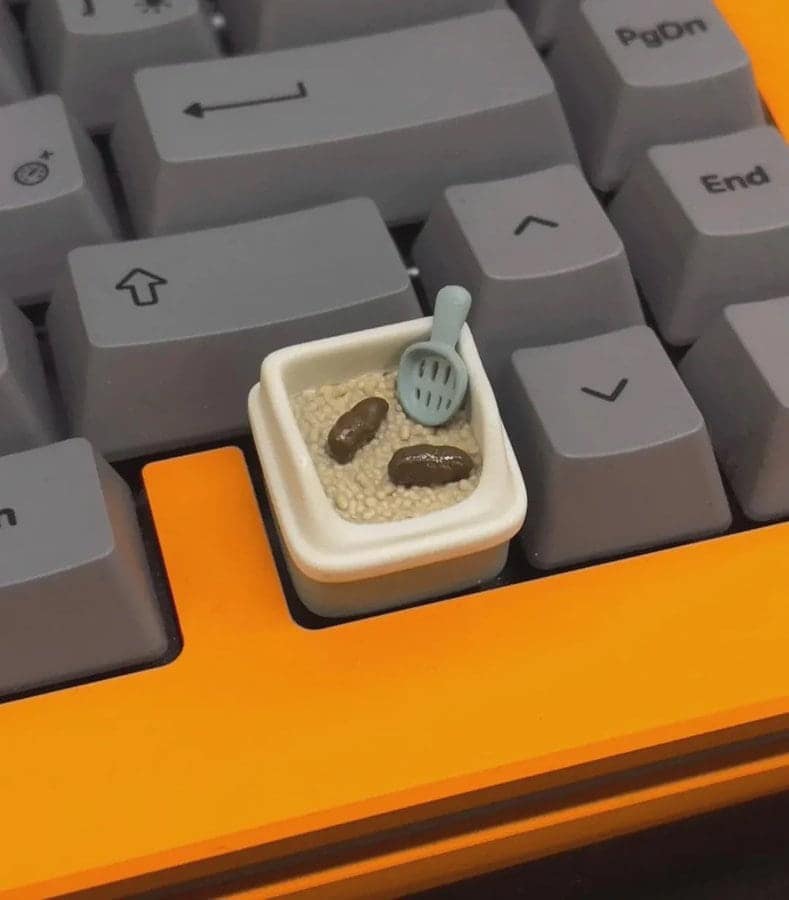A custom mechanical keyboard featuring an "artisan" keycap that is a hyper-realistic miniature replica of a cat's litter box, complete with tiny waste clumps and a plastic scooper.