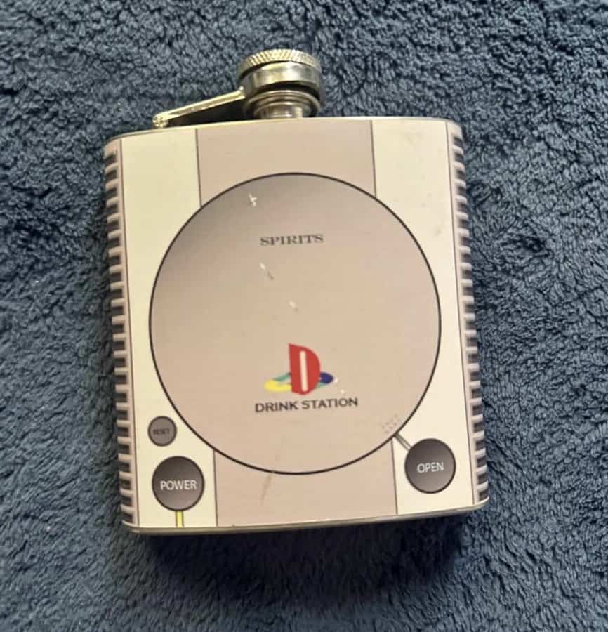 A stainless steel hip flask wrapped in a skin that perfectly replicates the original Sony PlayStation console, humorously rebranded as a "Spirits Drink Station."