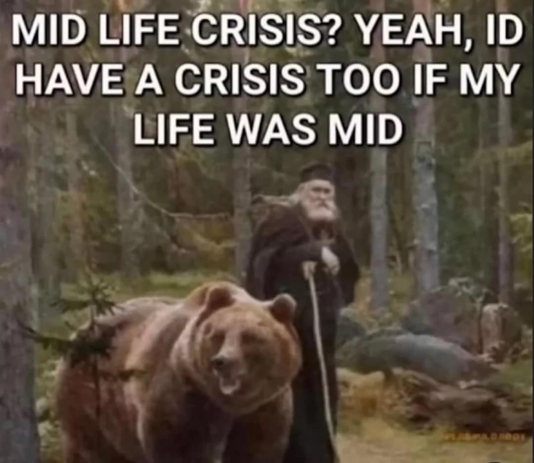 those classic memes featuring an old-school aesthetic: an elderly, bearded sage with a staff stands calmly next to a giant grizzly bear in a dense forest. The bold impact font text reads: "MID LIFE CRISIS? YEAH, ID HAVE A CRISIS TOO IF MY LIFE WAS MID."