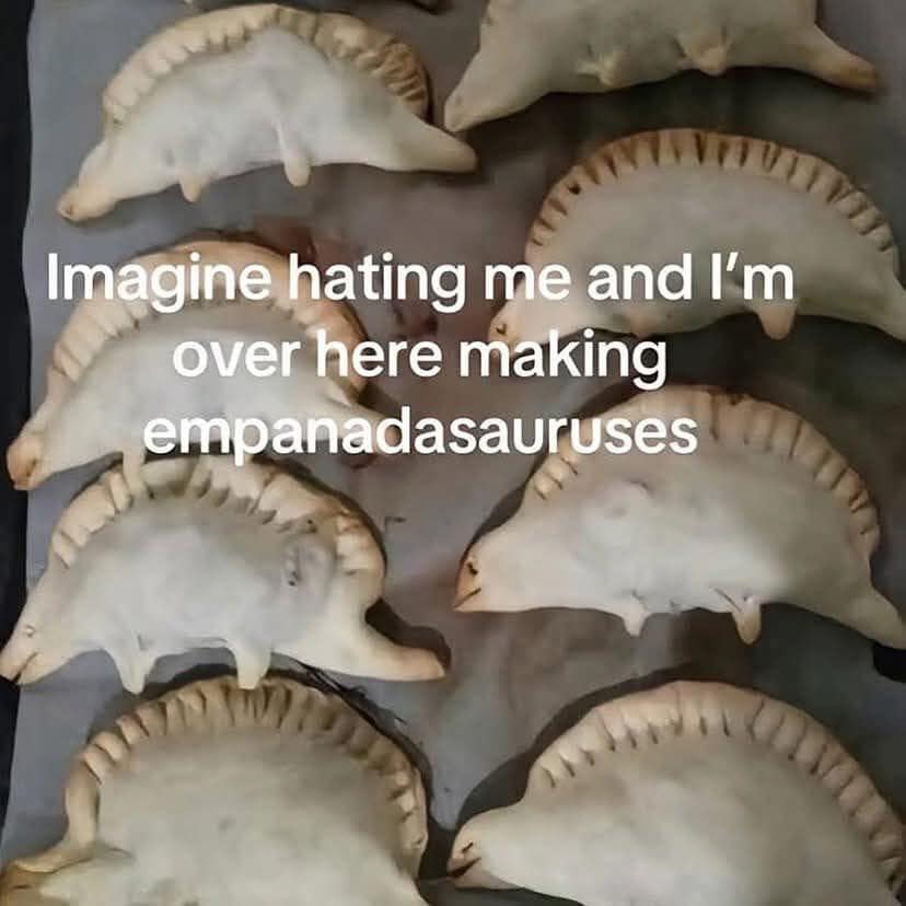 whimsical classic meme showing a baking tray full of "empanadasauruses"—empanadas carefully shaped to look like small dinosaurs with ridged backs and tiny legs. The caption reads: "Imagine hating me and I'm over here making empanadasauruses".