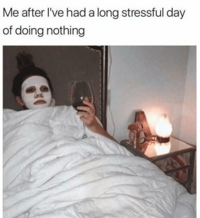 A quintessential introvert meme showing a person lying in bed tucked under a white duvet, wearing a white facial mask and holding a large glass of red wine. The text reads: "Me after I've had a long stressful day of doing nothing."