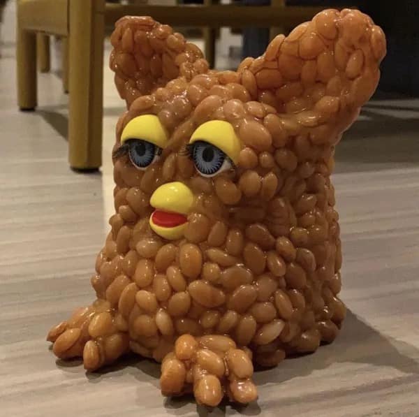 A deeply "cursed" funny meme of a 90s Furby toy whose fuzzy exterior has been completely replaced by a thick, wet layer of baked beans, creating a "Beany Furby" that is equal parts impressive and horrifying.
