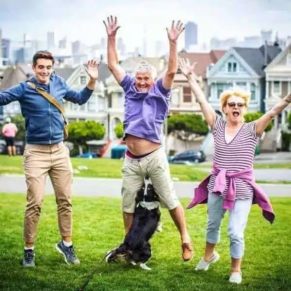 chaotic perfectly timed animal photo of three people jumping for joy in a park. A black and white dog is caught mid-leap, its snout perfectly positioned for an accidental "nutcracker" collision with the man in the center.