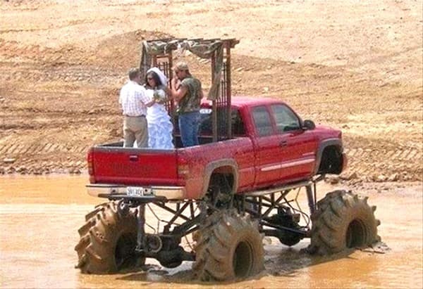 The ultimate rural wedding: a bride and groom exchange vows under a makeshift archway in the bed of a massive, red monster truck parked in the middle of a muddy off-road pit.