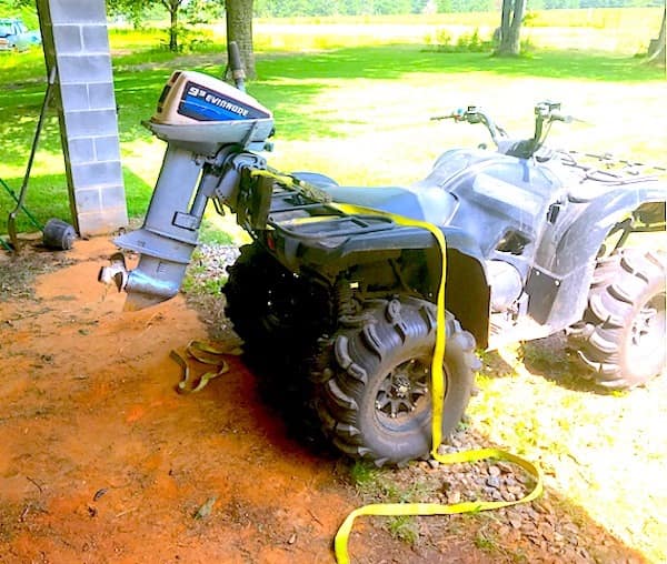 A bizarre redneck DIY car hybrid featuring a black utility ATV with an entire Evinrude boat outboard motor strapped to the rear rack with yellow webbing, propeller and all.