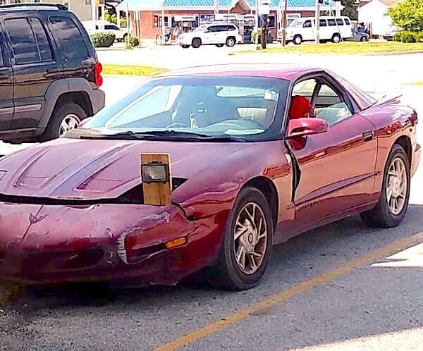 A low-budget repair for a maroon Pontiac Firebird. Instead of fixing the pop-up headlight, the owner bolted a 2x4 wooden plank into the empty socket to mount a small, rectangular off-road fog light.
