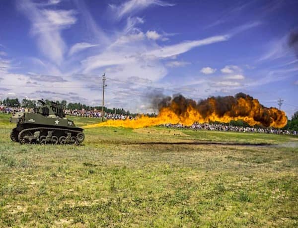 A high-intensity photo of a vintage military flamethrower tank unleashing a massive, roaring stream of fire across a field during a historical demonstration or mud-bogging event.