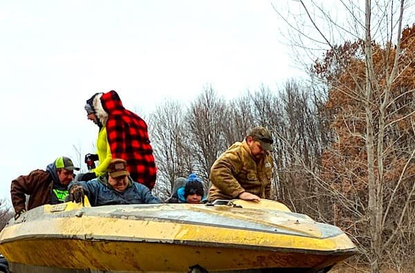 The "Land Yacht" in action: a yellow speedboat hull that has been professionally (or unprofessionally) mounted onto a high-clearance truck frame, carrying a full crew of people through a wooded area.