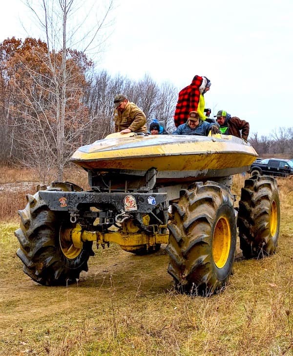 A front-facing view of the yellow "Boat Truck," showing the incredible scale of the massive, yellow-rimmed tractor tires supporting a vintage fiberglass speedboat hull as its primary cabin.