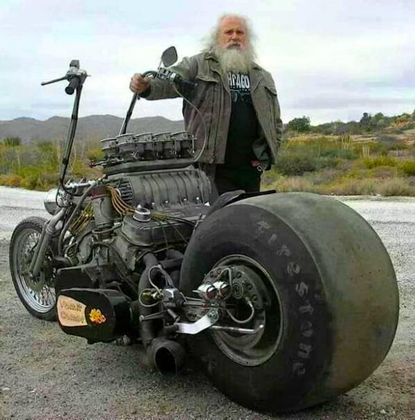 legendary redneck DIY car creation in motorcycle form. An elderly man with a long white beard stands next to a monstrous custom bike powered by a massive V8 engine with a towering blower. The rear is dominated by a single, colossal Firestone drag slick tire that is wider than the man himself.