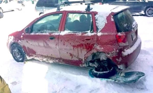 A winter survival hack showing a red hatchback missing its rear passenger-side wheel. The car is being driven through the snow with the rear hub resting directly inside a blue plastic snow sled to act as a ski.