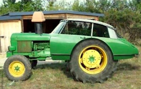 A surreal redneck DIY car hybrid featuring the front half of a green John Deere tractor seamlessly welded to the rear half of a vintage sedan. The vehicle retains the tractor’s massive rear tires and high-sitting cabin, complete with a custom chimney exhaust.
