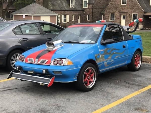 redneck DIY car build based on a blue Geo Metro. It features a massive hood scoop, red racing stripes, flame decals, a literal spoiler made from a bent metal pipe with lights, and a front splitter held on by hope and bolts.