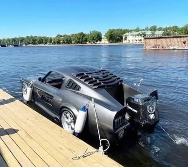 redneck DIY car conversion showing a classic grey Ford Mustang that has been turned into a fully functional speedboat. It is docked at a pier, featuring a chopped rear deck and a massive Mercury outboard motor mounted to the trunk area.