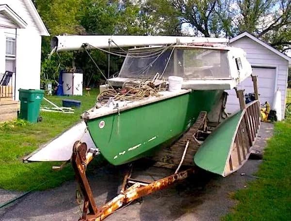 A confused redneck DIY car project where a green sailboat hull has been mounted onto a rusted trailer and modified with a wooden cabin structure. It features a mast laying across the top and appears to be a "land boat" parked in a residential yard.